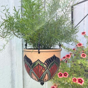 May include: A hanging ceramic planter with a speckled beige finish and a decorative carved pattern featuring red and dark green petal-like shapes. The bowl-shaped pot is suspended by black cords and contains a lush, feathery green asparagus fern.