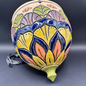 May include: A hanging ceramic planter with a teardrop shape, featuring a speckled beige base and a vibrant hand-painted floral pattern in shades of yellow, deep blue, green, and orange. The piece includes a grey cord for suspension.