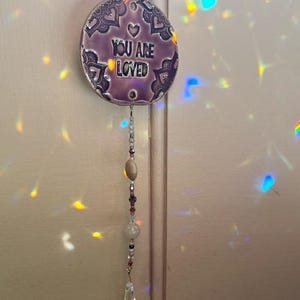 May include: A circular purple ceramic ornament featuring the embossed text YOU ARE LOVED and a heart symbol, surrounded by intricate dark patterns. A vertical strand of assorted beads and a clear crystal prism hangs from the bottom of the disc.