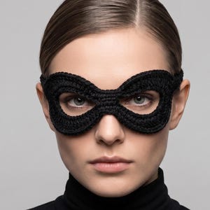 May include: A black crochet eye mask featuring a continuous, figure-eight shaped design that covers the eye area. The mask is constructed from dark yarn with a visible textured stitch pattern, secured around the head with a thin matching band.