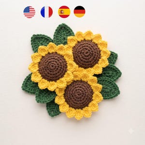 Könnte beinhalten: Eine handgefertigte Häkelapplikation bestehend aus drei Sonnenblumen mit dunkelbraunen Mitten und leuchtend gelben Blütenblättern, umgeben von mehreren dunkelgrünen gehäkelten Blättern. Das Arrangement ist kreisförmig und flach mit detaillierten Maschen.