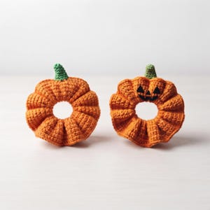 May include: Two orange crochet pumpkin hair scrunchies with green stems. One scrunchie is plain, while the other features a black stitched jack-o'-lantern face with triangular eyes and a jagged mouth. Both items have a circular, ruffled design.