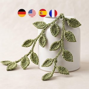 Może przedstawiać: Szydełkowa girlanda z wieloma szałwiowo-zielonymi liśćmi przymocowanymi do centralnej łodygi. Girlanda jest przewieszona przez gładki, cylindryczny biały wazon ceramiczny. Liście mają widoczną fakturę ściegów oraz spiczasty, owalny kształt.