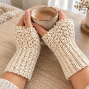 May include: A pair of cream-colored fingerless gloves featuring a textured, raised stitch pattern on the upper section and a vertical ribbed knit design on the wrist area. The gloves are crafted from a soft yarn and provide coverage for the hands.
