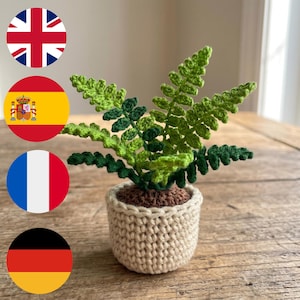 Puede incluir: Una planta de helecho tejida a ganchillo con hojas de color verde claro y oscuro, plantada en una base de tierra marrón texturizada y una maceta cilíndrica de color crema. La planta presenta múltiples frondas con detalles finos.