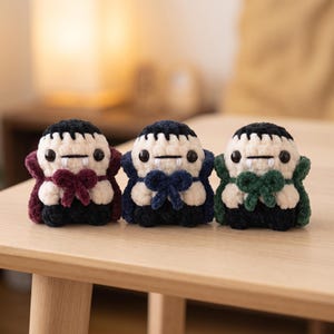 May include: Three small, handmade crochet vampire plushies featuring cream-colored faces, black hair, and two white fangs. Each plushie wears a cape and a bow in a different color: one in deep burgundy, one in navy blue, and one in dark forest green.