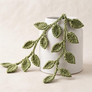 May include: A white ceramic vase draped with a long, crocheted vine featuring multiple sage green leaves. The vine is crafted with detailed stitching, showing individual leaf shapes attached to a central cord, resting against the smooth surface.