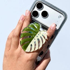 May include: A smartphone with a clear protective case and a triple-lens camera system, featuring a decorative phone grip shaped like a variegated monstera leaf with green and cream-colored segments attached to the back of the device.