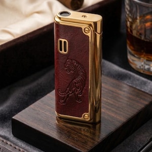 Peut inclure: Briquet doré avec un panneau en cuir bordeaux orné d'un motif de tigre. Le briquet est posé sur un socle en bois foncé. Un cigare, un verre de liquide ambré, un journal et un stylo sont également visibles.