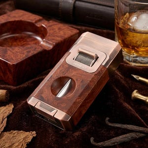 Peut inclure: Un coupe-cigare et briquet rectangulaire combiné, doté d'un cadre en métal cuivré poli et d'un corps au motif de grain de bois brun. L'appareil comprend une ouverture de lame circulaire, une molette d'allumage texturée et le mot HONEST.