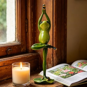 Puede incluir: Figura de rana verde brillante en pose de yoga, con los brazos levantados y una pierna extendida. La rana tiene ojos negros y detalles dorados. Una vela encendida en un vaso y un libro abierto con imágenes florales están cerca.