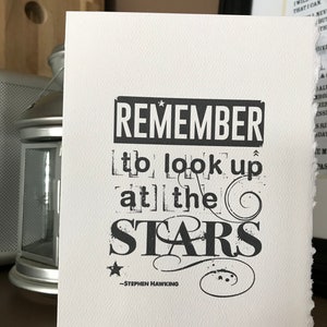 Puede incluir: Impresión tipográfica en blanco y negro con la cita "Recuerda mirar las estrellas" atribuida a Stephen Hawking.