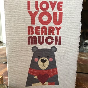 May include: A white card with red and pink text that reads "I love you beary much". A cartoon black bear with a red and black plaid scarf is holding a red heart.