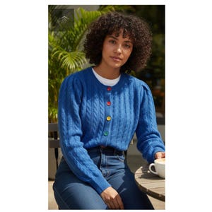 May include: A blue cable-knit cardigan with colorful buttons. The cardigan has a round neck and long sleeves. The buttons are red, orange, yellow, and green. The person is wearing a white shirt underneath and blue jeans.
