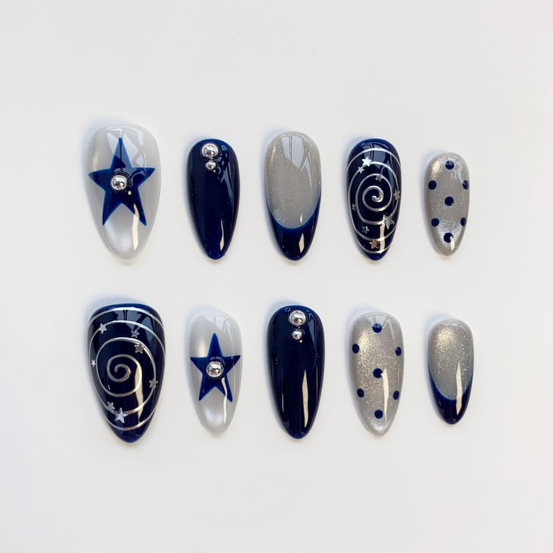 May include: A set of ten almond-shaped press-on nails featuring a navy blue and silver color palette. Designs include solid navy blue, silver glitter with navy tips, navy blue stars, silver swirls with tiny stars, and navy blue polka dots.