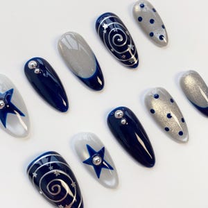 May include: A set of ten almond-shaped press-on nails featuring a color palette of navy blue and shimmering silver. Designs include solid navy blue with silver studs, silver glitter with navy blue polka dots, navy blue swirls with stars, and silver.