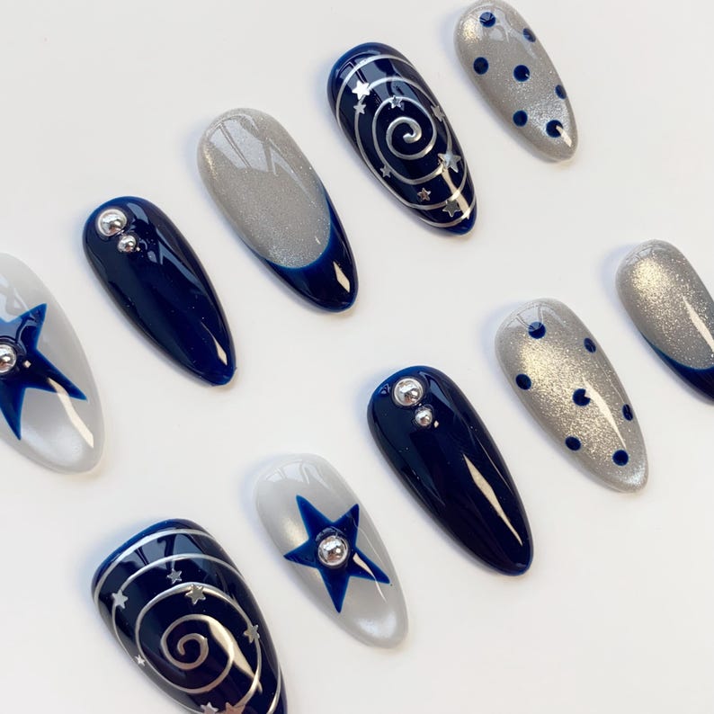 May include: A set of ten almond-shaped press-on nails featuring a color palette of navy blue and shimmering silver. Designs include solid navy blue with silver studs, silver glitter with navy blue polka dots, navy blue swirls with stars, and silver.