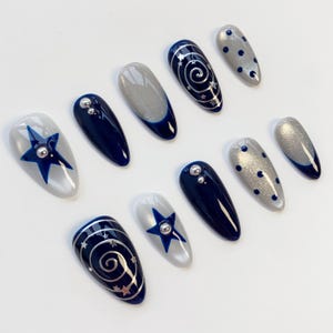 May include: A set of ten almond-shaped press-on nails featuring a navy blue and silver color palette. Designs include solid navy blue, silver glitter, navy blue stars, silver swirls with tiny star accents, and navy blue polka dots on silver.