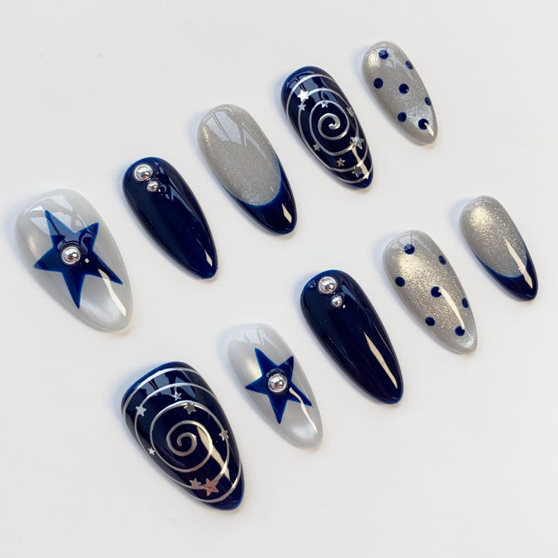 May include: A set of ten almond-shaped press-on nails featuring a navy blue and silver color palette. Designs include solid navy blue, silver glitter, navy blue stars, silver swirls with tiny star accents, and navy blue polka dots on silver.