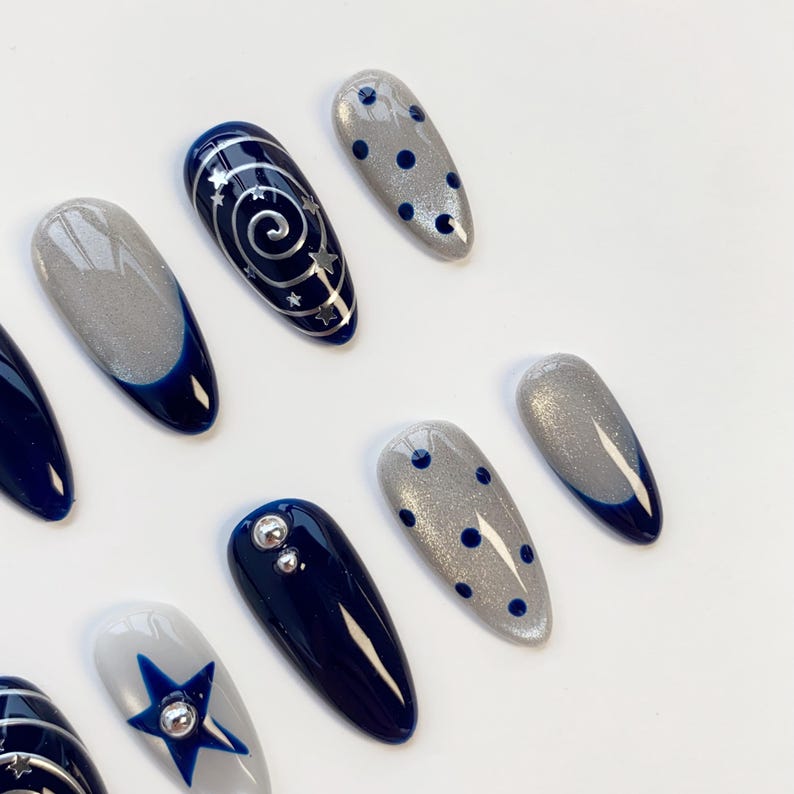 May include: A set of almond-shaped press-on nails featuring a dark navy blue and shimmering silver color palette. Designs include silver spiral and star patterns, dark blue polka dots on silver glitter, solid navy blue, and a blue star accent.