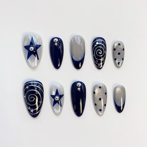 May include: A set of ten almond-shaped press-on nails featuring a color palette of navy blue, silver, and white. The designs include navy blue stars on white, solid navy blue with silver studs, silver glitter tips, silver spiral swirls, and navy blue dots.