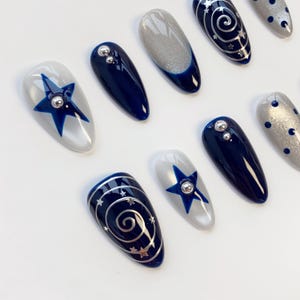 May include: A set of almond-shaped press-on nails featuring a celestial theme with deep navy blue and shimmering silver polish. Designs include blue stars with silver studs, silver spiral galaxies with tiny stars, and silver dots on a grey base.