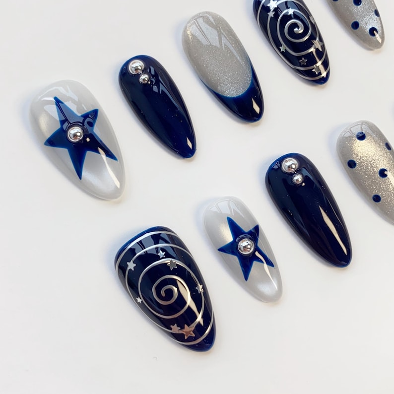 May include: A set of almond-shaped press-on nails featuring a celestial theme with deep navy blue and shimmering silver polish. Designs include blue stars with silver studs, silver spiral galaxies with tiny stars, and silver dots on a grey base.