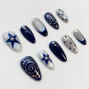 May include: A set of ten almond-shaped press-on nails featuring a color palette of navy blue, silver, and white. Designs include solid navy blue with silver studs, silver glitter with navy blue polka dots, navy blue swirls with stars, and blue stars.