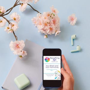 May include: A smartphone displays the cover of a book titled "Your Brain and Your Hormones" with a colorful logo. The image includes a light blue notebook, a mint-colored square, and light pink cherry blossoms on a light blue background.