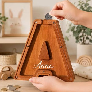 Può includere: Un salvadanaio in legno a forma di lettera A con un pannello frontale trasparente. Il legno ha una finitura marrone medio e il nome Anna è stampato in corsivo bianco sul davanti. Una fessura è posizionata in alto per inserire le monete.