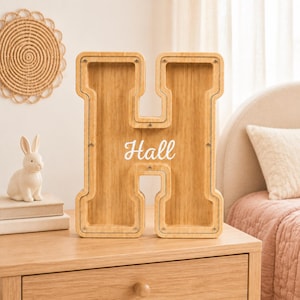 Può includere: Un salvadanaio in legno chiaro a forma di lettera H con un pannello frontale in acrilico trasparente. La parola Hall è stampata in corsivo bianco al centro del legno. La cornice è fissata con piccole viti metalliche sui bordi.
