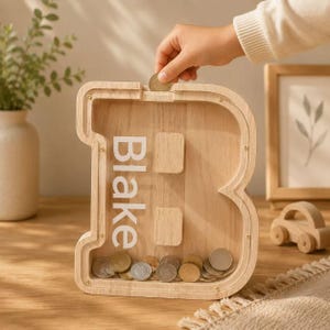 Può includere: Un salvadanaio in legno chiaro a forma di lettera B con un pannello frontale trasparente. Il nome Blake è stampato in bianco sulla parte anteriore. Una mano inserisce una moneta nella fessura superiore e diverse monete sono visibili sul fondo.