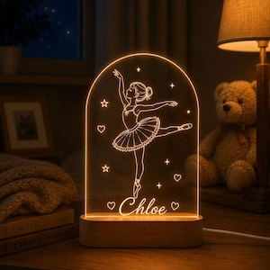 Op de afbeelding: Een boogvormig acryl nachtlampje met een warme oranje gloed, voorzien van een gegraveerde ballerina in een tutu omringd door sterren en hartjes. De voet is gemaakt van lichtgekleurd hout en de naam Chloe staat in cursief onderaan.