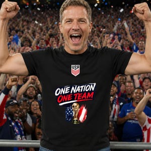 May include: A black short-sleeved t-shirt featuring a white USA crest logo at the top, the text "ONE NATION ONE TEAM" in white and red, and a graphic of the FIFA World Cup trophy flanked by stylized American flag and red-white-blue brush strokes.