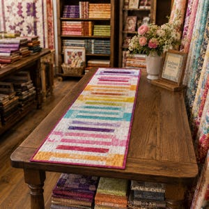 May include: A rectangular quilted table runner featuring a vibrant, multi-coloured horizontal striped pattern in shades of pink, orange, yellow, teal, and purple, finished with a solid dark pink border, displayed flat on a wooden table surface.