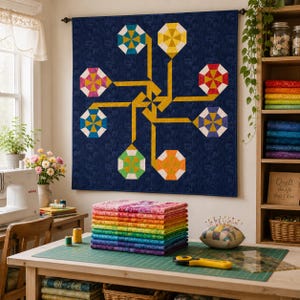Può includere: Una trapunta da parete con sfondo blu scuro e un motivo geometrico centrale di linee gialle che collegano sei forme ottagonali di vari colori. Una pila di tessuti piegati color arcobaleno è appoggiata su un tavolo sotto la trapunta.