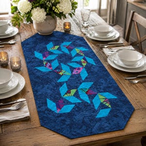 May include: A rectangular table runner featuring a dark blue mottled fabric base with a central decorative pattern of bright cyan geometric shapes. The design includes small, colorful, multi-patterned accents within the cyan elements, creating a repeating motif along the center of the runner.