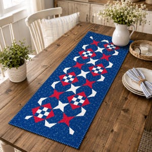 May include: A rectangular table runner featuring a dark blue fabric base with a speckled texture. The runner displays a repeating quilt pattern of red squares, white four-pointed stars, and small blue and white checkered square blocks.