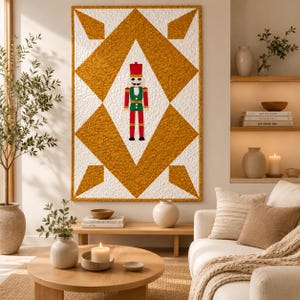 May include: A rectangular wall quilt featuring a large central diamond pattern in textured mustard yellow fabric against a white background, with a small, centered nutcracker figure wearing a red, green, and white uniform with a tall hat.