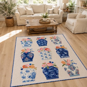 May include: A rectangular quilt featuring a cream-colored background with a repeating pattern of nine blue and white ceramic vases, each filled with colorful floral arrangements of pink, orange, and yellow flowers, finished with a blue border.