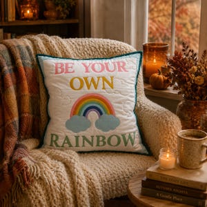 May include: A square white quilted throw pillow featuring the embroidered text BE YOUR OWN RAINBOW in pink, yellow, and green. The design includes a colorful rainbow arching over two light blue clouds, all finished with a dark teal border.