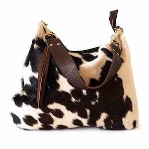 May include: A hobo-style handbag featuring a black and white cowhide pattern with a textured surface. The bag includes a dark brown leather strap attached with gold-toned metal hardware, including swivel clips and rivets on each side.