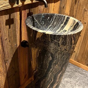 Peut inclure: Un lavabo sur pied autoportant en pierre marbrée noire et beige de forme conique, associé à un robinet mural noir mat doté de deux poignées en croix et d'un bec horizontal s'étendant au-dessus de la vasque.