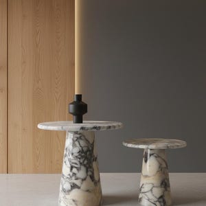 Peut inclure: Deux tables d'appoint en marbre blanc veiné de gris, chacune dotée d'un plateau circulaire et d'une base conique effilée. Un petit vase géométrique noir mat est posé au centre de la surface de la table la plus grande, créant un contraste.
