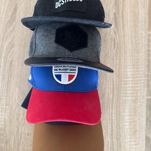 Può includere: Una pila di cinque cappellini da baseball in vari colori. Il cappellino superiore è nero con "DCSHOECO" in bianco. Sotto c'è un cappellino grigio con una toppa nera. Il terzo cappellino è blu e rosso con un emblema bianco. Il cappellino inferiore è marrone.