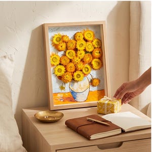 May include: A framed 3D wall art piece featuring a collection of bright yellow dried strawflowers arranged to mimic a vase of sunflowers. The artwork is encased in a light-colored wooden frame, showcasing textured floral elements that extend outward.