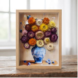 Könnte beinhalten: Ein rechteckiger Schattenfugenrahmen aus hellem Holz mit einem Druck einer blau-weißen Vase im Mosaikstil, gefüllt mit einem lebendigen Strauß aus getrockneten Strohblumen in Orange, Gelb, Violett und Creme, mit verstreuten Blütenblättern.