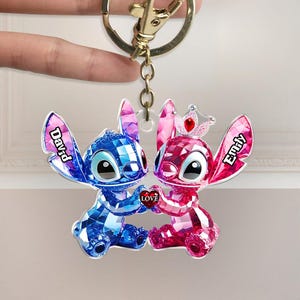May include: A personalised keyring featuring two cartoon characters, one blue and one pink, holding a heart that says "Love." The blue character has the name "David" on its ear, and the pink character has "Emily." The keyring is attached to a gold-coloured clasp.