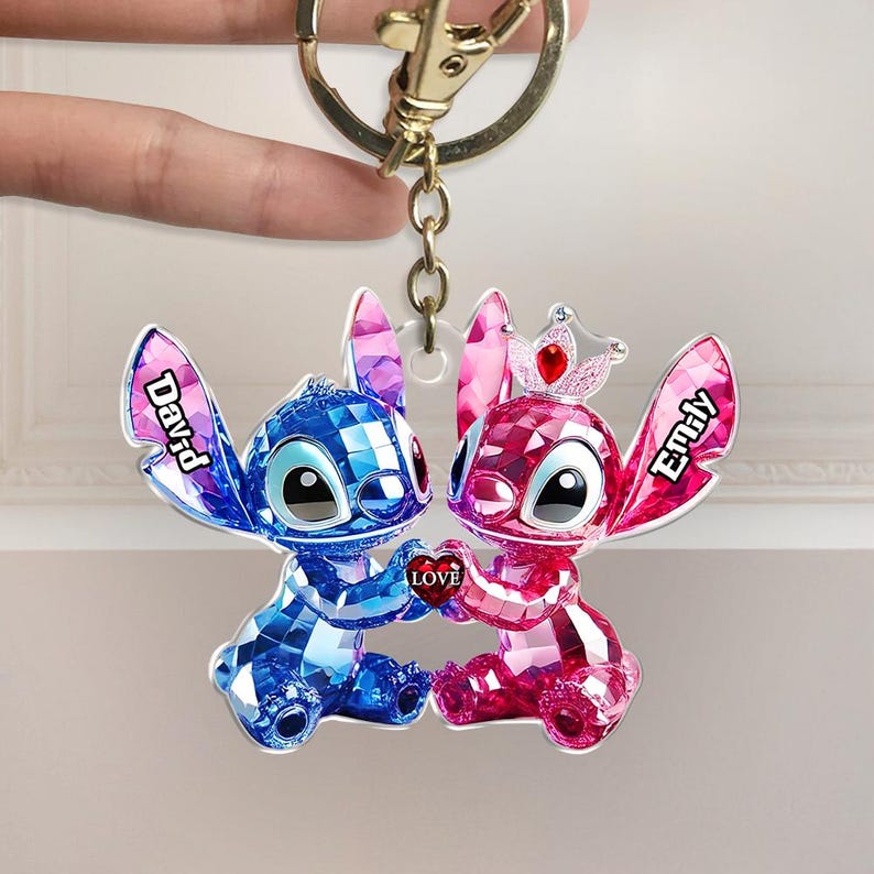 May include: A personalised keyring featuring two cartoon characters, one blue and one pink, holding a heart that says "Love." The blue character has the name "David" on its ear, and the pink character has "Emily." The keyring is attached to a gold-coloured clasp.