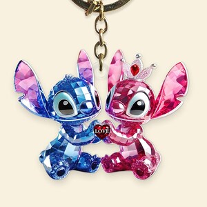 May include: A keychain with two crystal-effect figures, one blue and one pink, holding a heart with the word "LOVE." The pink figure wears a crown. The figures are connected by a gold-coloured chain.